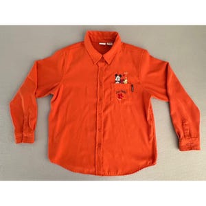 May include: Bright orange button-down shirt with long sleeves. The shirt features a small pocket on the chest with an embroidered design of Mickey Mouse and a reindeer. The text "Just Chillin'" is embroidered below the design. The shirt has a collared neckline.