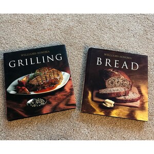 May include: Two Williams-Sonoma hardcover cookbooks. The left book is titled Grilling and features a grilled steak on a white plate. The right book is titled Bread and shows a loaf of sliced bread with butter on a wooden board.