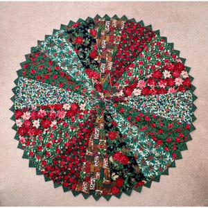 May include: A circular fabric tree skirt featuring a patchwork design of various holiday-themed patterns. The segments display red poinsettias, green holly leaves, and plaid motifs in a starburst arrangement with a scalloped, zigzag edge.