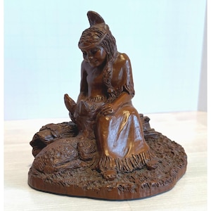 Könnte beinhalten: Braune Skulptur einer sitzenden indianischen Frau, die nach unten blickt, mit einem Bison. Die Frau trägt ein Kleid mit Fransen und hat lange geflochtene Haare. Die Skulptur steht auf einem Sockel, der Gras und Holz ähnelt.