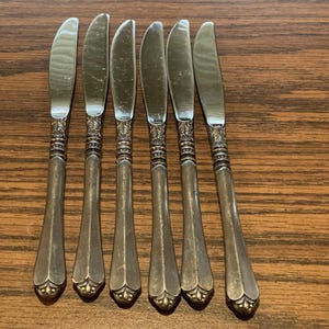 May include: A set of six vintage silver butter knives. Each knife has an ornate handle and a curved blade. The knives are arranged in a row on a wooden surface. The handles feature a detailed design, and the blades are polished.
