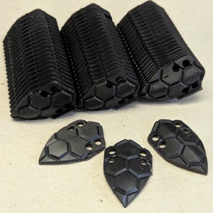 May include: An assortment of black, geometric-patterned components. Three cylindrical pieces with ridged sides and hexagonal ends are arranged above three shield-shaped pieces with a similar pattern. The items are made of a matte black material.