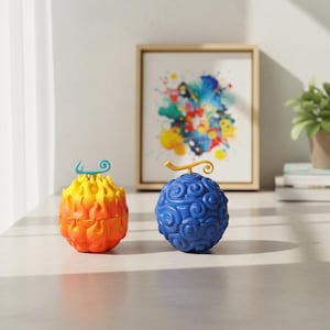 May include: Two colorful, stylized fruit-shaped objects. One is orange and yellow, resembling flames, and the other is blue with a swirling pattern. They are placed on a light-colored surface, with a framed abstract painting and a potted plant in the background.