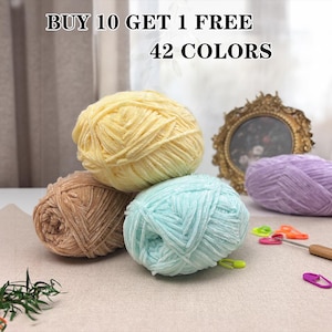 Op de afbeelding: Vier bollen garen in pastelkleuren: geel, lichtbruin, blauwgroen en lavendel. De afbeelding bevat ook een haaknaald, steekmarkeerders en een decoratief gouden frame. De tekst bovenaan luidt "BUY 10 GET 1 FREE 42 COLORS."