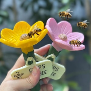Puede incluir: Dos adornos con forma de flor, uno amarillo y otro rosa, cada uno con una abeja encima. Los objetos tienen tallos verdes y pequeños letreros con texto. El fondo está borroso, lo que sugiere un entorno al aire libre.