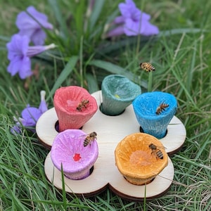 May include: A wooden flower-shaped bee waterer with five colorful cups. Each cup is a different color: red, green, blue, purple, and orange. Bees are drinking from the water in the cups. The waterer is set on green grass with purple flowers in the background.