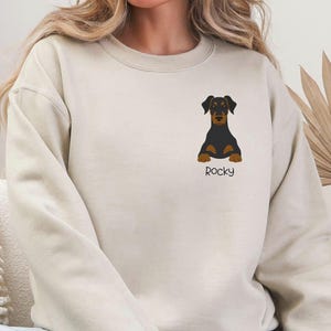 May include: A beige sweatshirt featuring a black and brown dog graphic with the name "Rocky" below it. The dog is centered on the chest. The sweatshirt is a casual style, suitable for everyday wear.