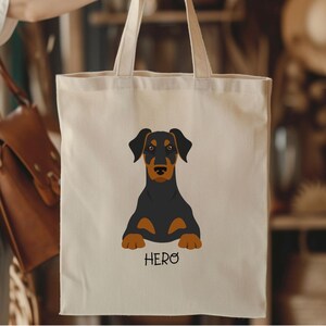 May include: Cream-colored tote bag featuring a black and brown Doberman Pinscher illustration. The dog is lying down with the word "HERO" printed below. The bag is held by a person.