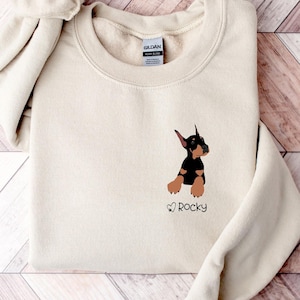 May include: A cream-colored sweatshirt featuring a small embroidered graphic of a Doberman Pinscher dog in black and brown. The dog's name, "Rocky," is printed below the graphic in a script font. The sweatshirt has a crew neck and long sleeves.