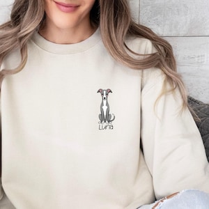 K&ouml;nnte beinhalten: Cremefarbenes Sweatshirt mit einer kleinen, gestickten Illustration eines Windhundes und dem Namen "Luna" darunter. Der Hund ist sitzend dargestellt. Das Sweatshirt hat eine neutrale Farbe.