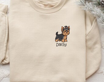 Könnte beinhalten: Ein beigefarbenes Sweatshirt mit Rundhalsausschnitt und einer gestickten Illustration eines Yorkshire Terriers auf der linken Brustseite. Unter dem Hund ist der Name Daisy in schwarzer Schreibschrift gestickt. Das Sweatshirt hat lange Ärmel.