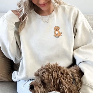 May include: Cream-colored sweatshirt featuring a cartoon dog graphic with the name "MIMI" below it. The dog is light brown with a white chest. The sweatshirt has a crew neck and long sleeves. The person is wearing blue jeans.