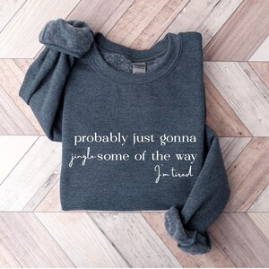 Puede incluir: Una sudadera gris oscuro con el texto "probably just gonna jingle some of the way I'm tired" en escritura blanca. La sudadera est&aacute; doblada sobre una superficie de madera con un patr&oacute;n de espiga.