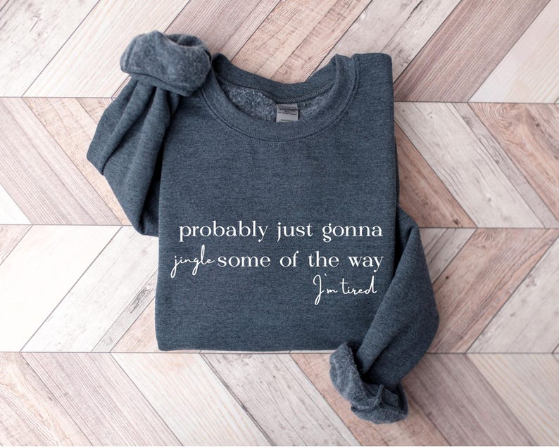 Puede incluir: Una sudadera gris oscuro con el texto "probably just gonna jingle some of the way I'm tired" en escritura blanca. La sudadera est&aacute; doblada sobre una superficie de madera con un patr&oacute;n de espiga.