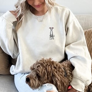 K&ouml;nnte beinhalten: Cremefarbenes Sweatshirt mit einer kleinen, gestickten Illustration eines Windhundes und dem Namen "Luna" darunter. Der Hund ist im Sitzen dargestellt. Das Sweatshirt hat einen Rundhalsausschnitt und lange &Auml;rmel.