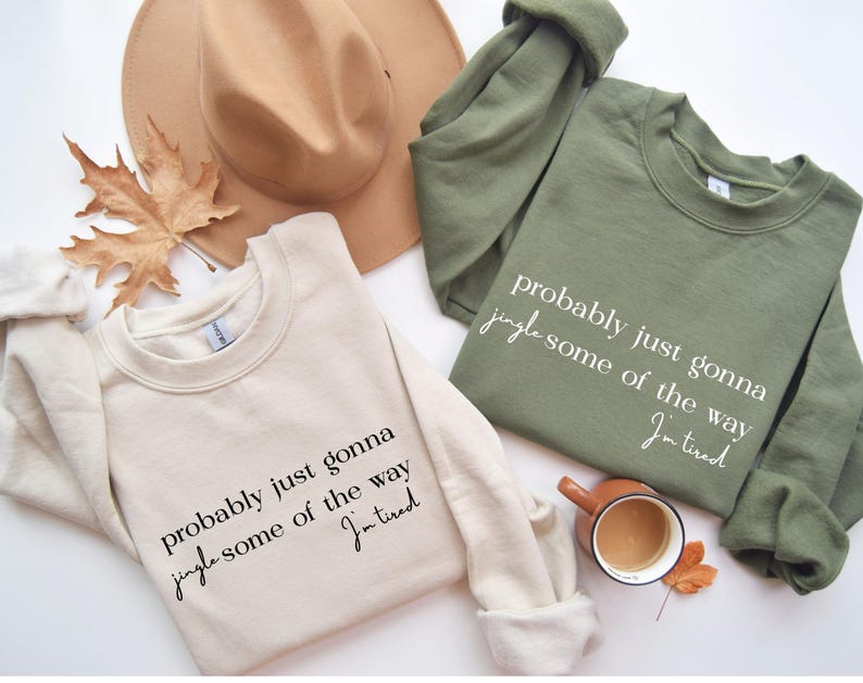 Puede incluir: Dos sudaderas, una crema y otra verde oliva, con el texto "probably just gonna jingle some of the way, I'm tired." Un sombrero marr&oacute;n y una hoja de oto&ntilde;o tambi&eacute;n est&aacute;n en la imagen. Una peque&ntilde;a taza de caf&eacute; est&aacute; al lado de la sudadera verde.