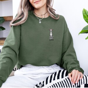 K&ouml;nnte beinhalten: Ein olivgr&uuml;nes Sweatshirt mit einer gestickten Illustration eines Windhundes und dem Namen "Luna". Der Hund hat ein rotes Halsband. Das Sweatshirt hat einen Rundhalsausschnitt und lange &Auml;rmel. Das Design befindet sich auf der linken Brust.