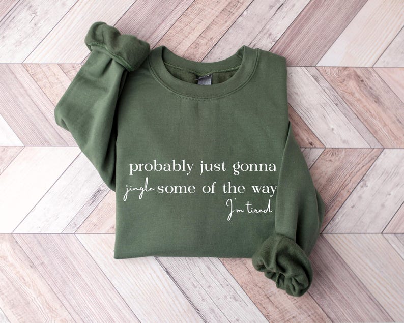 Puede incluir: Una sudadera verde oliva con el texto "probably just gonna jingle some of the way, I'm tired" en escritura blanca. La sudadera est&aacute; doblada sobre una superficie de madera marr&oacute;n claro y blanca.