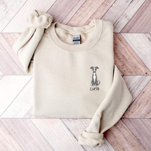 K&ouml;nnte beinhalten: Ein cremefarbenes Sweatshirt mit Rundhalsausschnitt und langen &Auml;rmeln. Das Sweatshirt zeigt eine gestickte Illustration eines Windhundes mit dem Namen "Luna" unter dem Hund. Das Sweatshirt ist auf einer Holzoberfl&auml;che gefaltet.