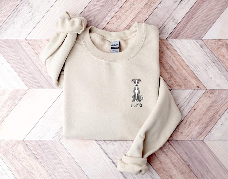 K&ouml;nnte beinhalten: Ein cremefarbenes Sweatshirt mit Rundhalsausschnitt und langen &Auml;rmeln. Das Sweatshirt zeigt eine gestickte Illustration eines Windhundes mit dem Namen "Luna" unter dem Hund. Das Sweatshirt ist auf einer Holzoberfl&auml;che gefaltet.