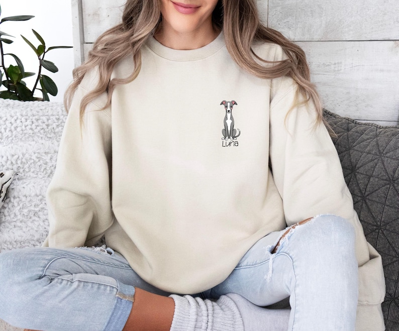 K&ouml;nnte beinhalten: Cremefarbenes Sweatshirt mit einer kleinen, gestickten Illustration eines Windhundes und dem Namen "Luna" darunter. Der Hund ist sitzend dargestellt. Das Sweatshirt hat eine neutrale Farbe.