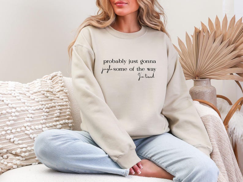 Puede incluir: Sudadera beige con el texto "probably just gonna jingle some of the way, I'm tired" en escritura negra. La sudadera se usa con vaqueros azul claro. La imagen es de una persona sentada en un sof&aacute;.