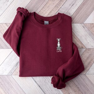 K&ouml;nnte beinhalten: Ein weinrotes Sweatshirt mit Rundhalsausschnitt. Es hat eine gestickte Illustration eines Windhundes und den Namen "Luna" in Schreibschrift. Das Sweatshirt ist auf einer hellen Holzmaserung gefaltet.