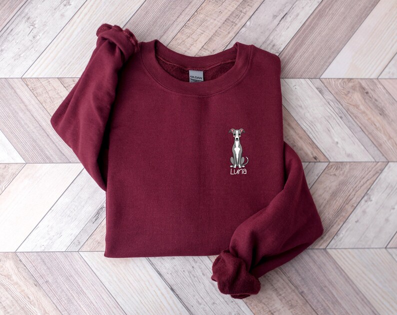 K&ouml;nnte beinhalten: Ein weinrotes Sweatshirt mit Rundhalsausschnitt. Es hat eine gestickte Illustration eines Windhundes und den Namen "Luna" in Schreibschrift. Das Sweatshirt ist auf einer hellen Holzmaserung gefaltet.