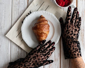 Puede incluir: Naturaleza muerta con un cruasán en un plato blanco, una taza de rosas rojas y un periódico. Las manos llevan guantes negros transparentes con estampado de leopardo. La escena se desarrolla sobre una superficie de madera blanca.