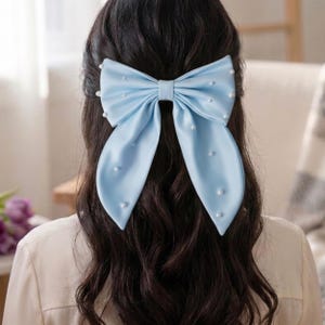 Può includere: Un fiocco per capelli in raso azzurro chiaro con dettagli di perle bianche. Il fiocco è legato a forma di fiocco grande ed è attaccato a capelli scuri. Un accessorio per capelli elegante.