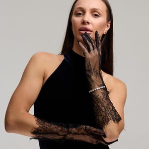 May include: A woman wearing black lace gloves with a floral design. The gloves extend to the elbows and are paired with a dark blue halter top dress. A silver bracelet with small stones is visible on the wrist.