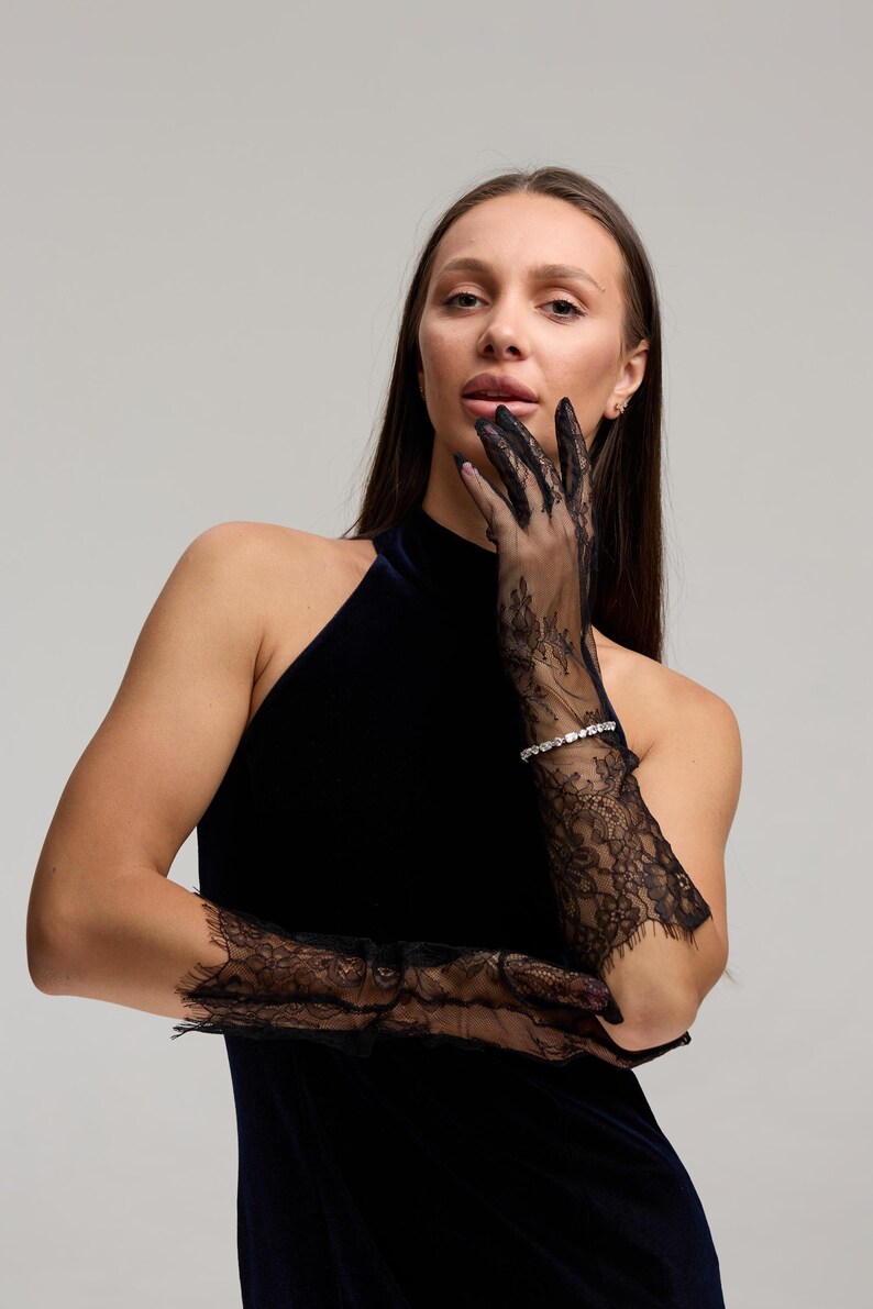 May include: A woman wearing black lace gloves with a floral design. The gloves extend to the elbows and are paired with a dark blue halter top dress. A silver bracelet with small stones is visible on the wrist.