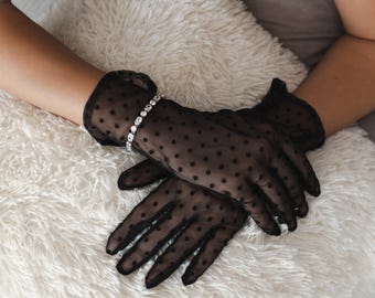 May include: Black sheer gloves with a polka dot pattern. A sparkling bracelet with clear stones adorns the wrist. The gloves are resting on a fluffy white surface, creating a contrast of textures.