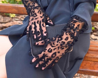 Puede incluir: Guantes negros transparentes con estampado de leopardo. Los guantes son largos, se extienden más allá de la muñeca y se combinan con un bolso de mano gris oscuro. Los guantes tienen un borde con volantes en la muñeca.