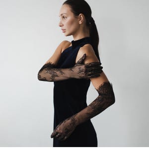以下が含まれることがあります： ノースリーブの黒いドレスと、長い黒のシースルーレースの手袋を着用した女性。手袋は複雑な花柄で、上腕まで伸びています。ドレスはハイネックです。