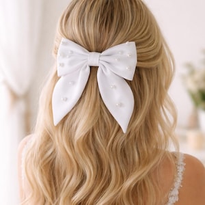 May include: A white satin hair bow adorned with small, scattered pearls. The bow is centered on blonde wavy hair, with the back of the head and shoulders visible. The bow's design features a classic bow shape with long, flowing tails.