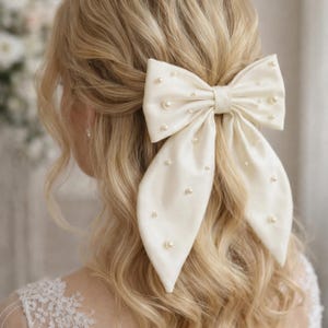 Puede incluir: Lazo de pelo de satén color marfil adornado con perlas de imitación dispersas. El lazo grande está peinado en el cabello, con largas colas fluidas. Este accesorio para el cabello añade un toque de elegancia y es adecuado para bodas u ocasiones especiales.