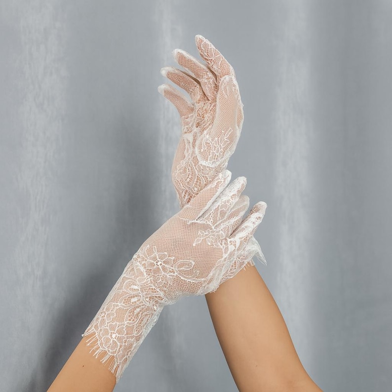 May include: A pair of white lace gloves. The gloves are made of a delicate, floral-patterned lace and are designed to cover the hands. The gloves are shown against a gray background.