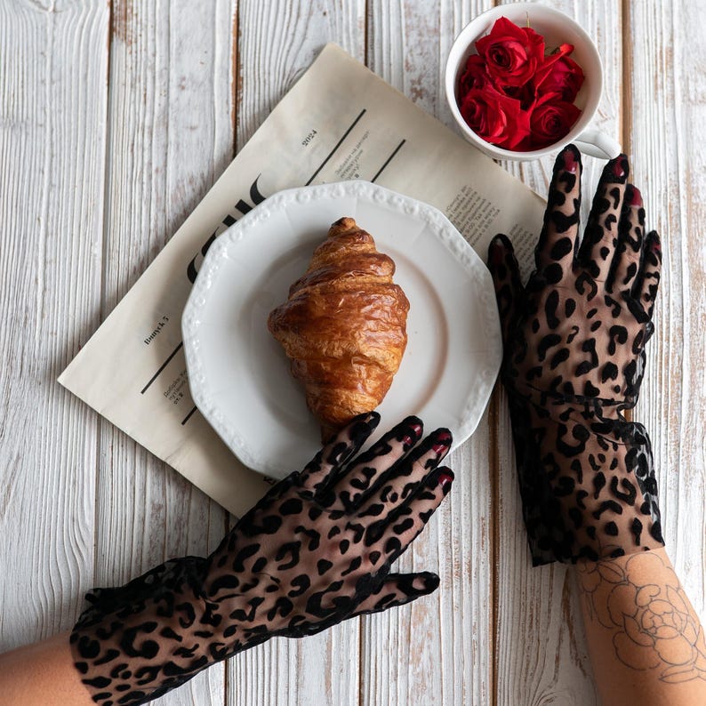 Pode incluir: Natureza morta com um croissant num prato branco, uma ch&aacute;vena de rosas vermelhas e um jornal. As m&atilde;os usam luvas pretas transparentes com estampa de leopardo. A cena &eacute; definida numa superf&iacute;cie de madeira branca.