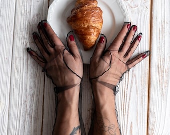 Può includere: Un croissant dorato su un piatto bianco. Il piatto è tenuto da mani con guanti neri trasparenti. Le unghie sono dipinte di rosso. Lo sfondo è una superficie di legno bianco.