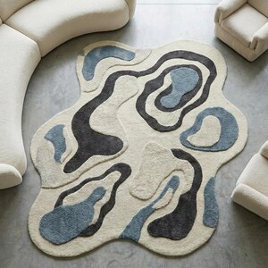 Può includere: Vista dall'alto di un moderno soggiorno. Un divano curvo color crema e due poltrone abbinate circondano un tappeto astratto unico con forme organiche blu e grigie. Il tappeto è posizionato su un pavimento in cemento grigio.