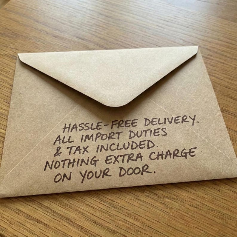 A brown kraft paper envelope resting on a light wooden table. Handwritten text in black marker on the envelope reads: "HASSLE-FREE DELIVERY. ALL IMPORT DUTIES & TAX INCLUDED. NOTHING EXTRA CHARGE ON YOUR DOOR."