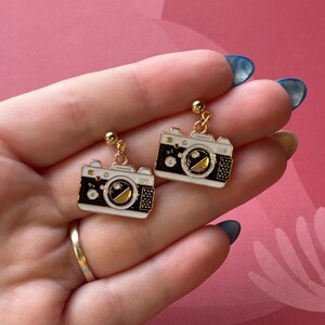 May include: A pair of gold-coloured earrings with a camera design. The earrings have a black and white body with gold accents and a small gold ball at the top. The earrings are suspended from a gold-coloured post.