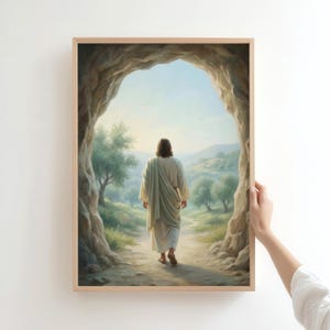 Könnte beinhalten: Ein gerahmter Wandkunstdruck, der eine langhaarige Gestalt in einem hellen Gewand zeigt, die von einem dunklen, felsigen Höhleneingang weg in eine helle, sonnendurchflutete Landschaft mit grünen Bäumen und sanften Hügeln in der Ferne geht.