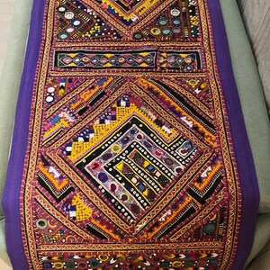 Könnte beinhalten: Ein langer, rechteckiger Tischläufer mit einem tiefvioletten Rand und aufwendigen Stickereien in Gelb, Orange, Schwarz und Weiß. Das Design umfasst geometrische Rautenmuster, kleine runde Spiegel und detaillierte Handarbeiten.