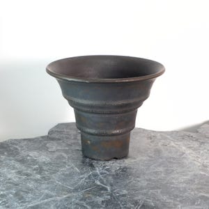 Puede incluir: Una maceta de cerámica gris oscuro, escalonada, con un borde ancho y acampanado. La maceta tiene un aspecto texturizado y envejecido y se asienta sobre una superficie de piedra gris. El diseño presenta crestas horizontales.