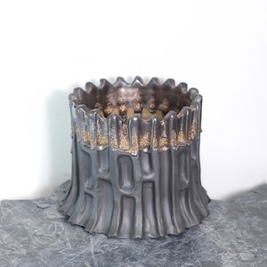 以下が含まれることがあります： 樹皮のような質感の、円筒形のグレーのセラミック花瓶。上端は波状のデザインで、金色のアクセントの帯があります。花瓶の内側は、より濃い茶色です。