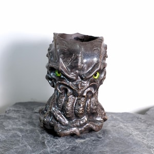 May include: A dark bronze-colored, sculpted vase with a monster-like face. The vase has a textured surface, with green eyes and a detailed, menacing expression. The vase is a unique decorative item.