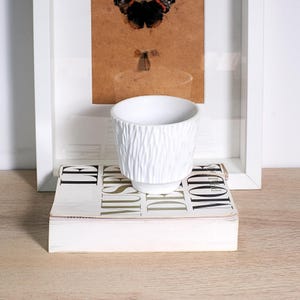 Peut inclure: Un pot de fleurs en céramique blanche avec un motif texturé en forme d'écorce. Le pot est posé sur un livre avec les mots "LE MUSEE DE MODE" imprimés sur la couverture. Une œuvre d'art encadrée avec une image de papillon est en arrière-plan.