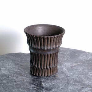 Puede incluir: Un jarrón de cerámica marrón oscuro con un diseño acanalado vertical texturizado. El jarrón tiene forma cilíndrica con un borde ligeramente ensanchado. El jarrón mide aproximadamente 13 cm de alto y está sobre una superficie de piedra gris.